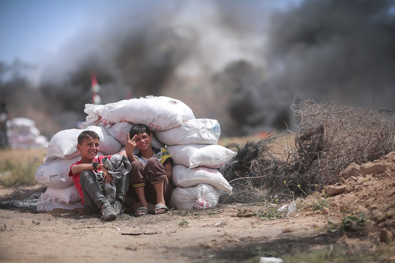 2 unarmed Palestinian boys sitting behind a pile of concrete bags in the battle field smiling. The boy on the right is making the victory / peace sign with his hand